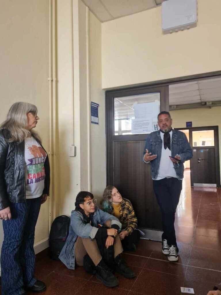 Selene Mira observa a su compañero, Ariel Sanabria, que está hablando. Ambos están de pie, en el medio, dos chicas están sentadas en el piso